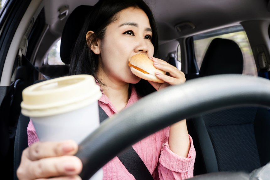 risky behavior driving concept with Asian woman eating burger and drinking coffee while driving a vehicle, poses accident risk and traffic crashes