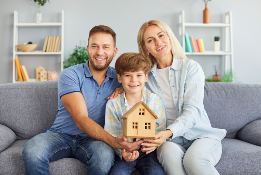 Portrait of happy joyful young family of father, mother and child boy holding wooden house in hands and looking cheerful at camera sitting on sofa at home. Real estate and mortgage concept.