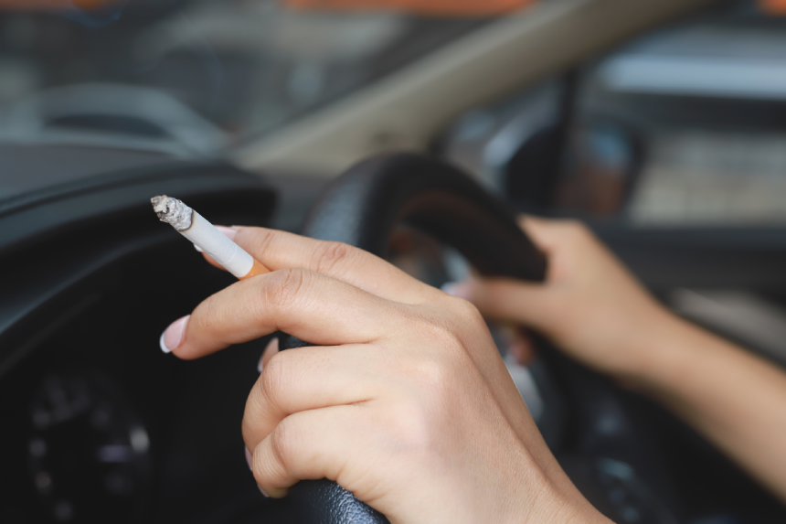 Hand holding burning cigarette while gripping steering wheel, with smoke filling car.