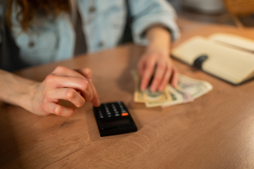 Woman's hands are busy calculating her expenses using a small black calculator, organizing a stack of dollar bills, and managing household finances for budgeting and paying utilities
