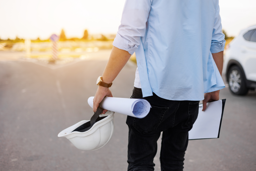 Unrecognisable engineer, architect or worker holds in hand protective white hard hat and blueprint on multi-apartment residential complex background, sunlighting.