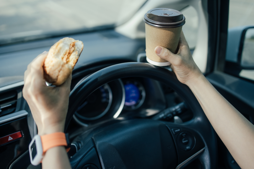 Woman driving car while holding coffee and eating hamburger