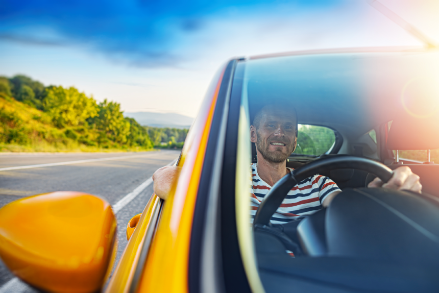 A happy middle-aged man smiles while driving a vibrant yellow car along a scenic country road during a sunny summer day.