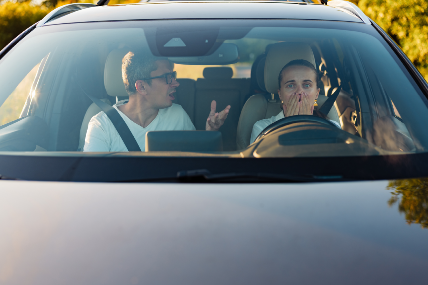 Two people sit inside a car, one is animatedly talking while the other looks shocked. They appear to be having a serious discussion on a bright afternoon drive.