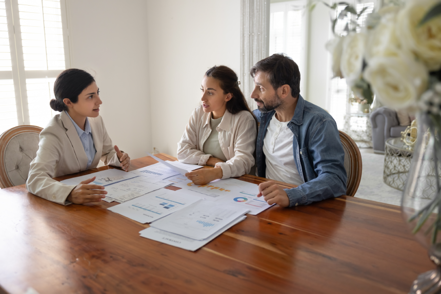 Professional consultant meets with couple at table covered with documents, graphs, and data charts, explains points, specific sections, review detailed plans or reports.