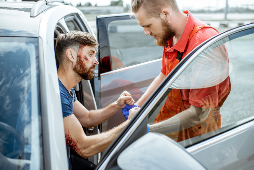Ambulance worker examining injuries of a man sitting near the car after the road accident, providing emergency medical assistance