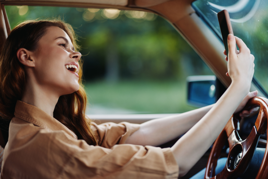 Young woman enjoying a joyful moment while driving in a vintage car, taking a selfie against a lush green background with natural light.