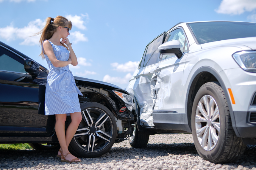 Sad young woman driver standing near her smashed car looking shocked on crashed vehicles in road accident