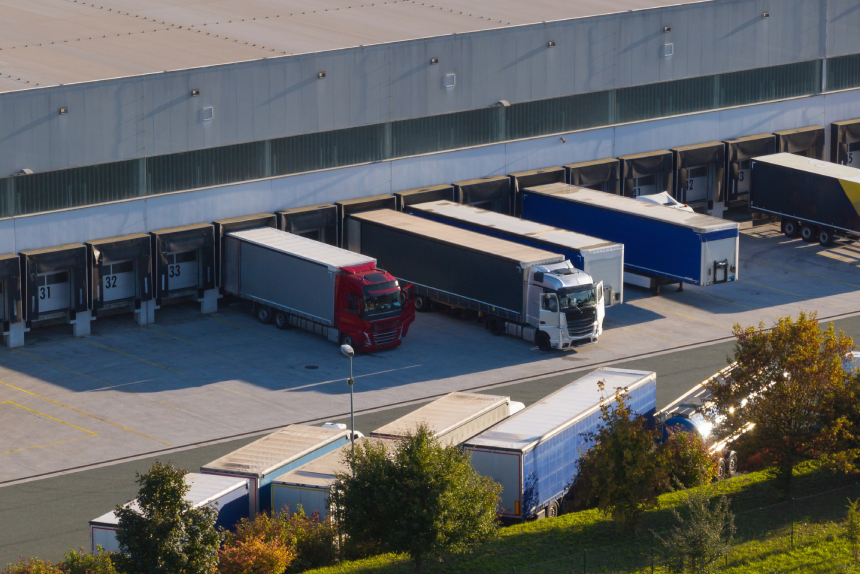 Trucks handle cargo at a large logistics center with loading docks and warehouse operations seen from above.
