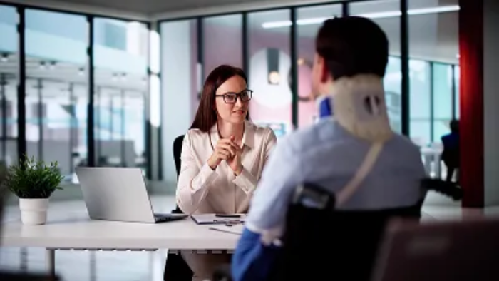 A professional woman in an office setting sitting across from a person in a wheelchair wearing a neck brace and arm sling.