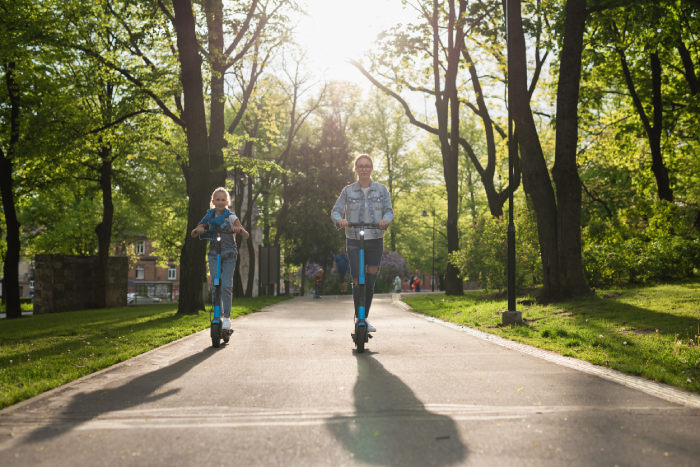 Happy mother and daughter riding electric scooters in city park